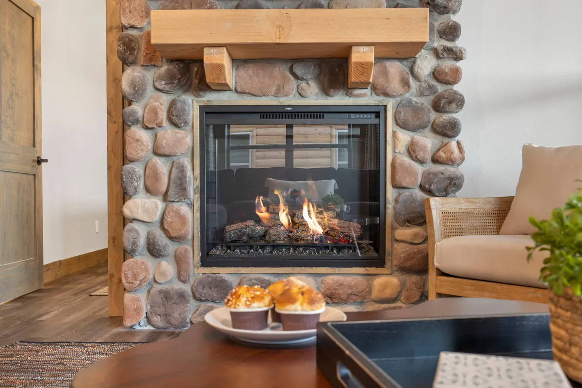 A welcoming great room featuring a cozy fireplace with a warm glow, a comfortable seating arrangement perfect for gatherings, and a display of muffins on the coffee table. The space is sunlit, highlighting the sense of openness and inviting atmosphere.