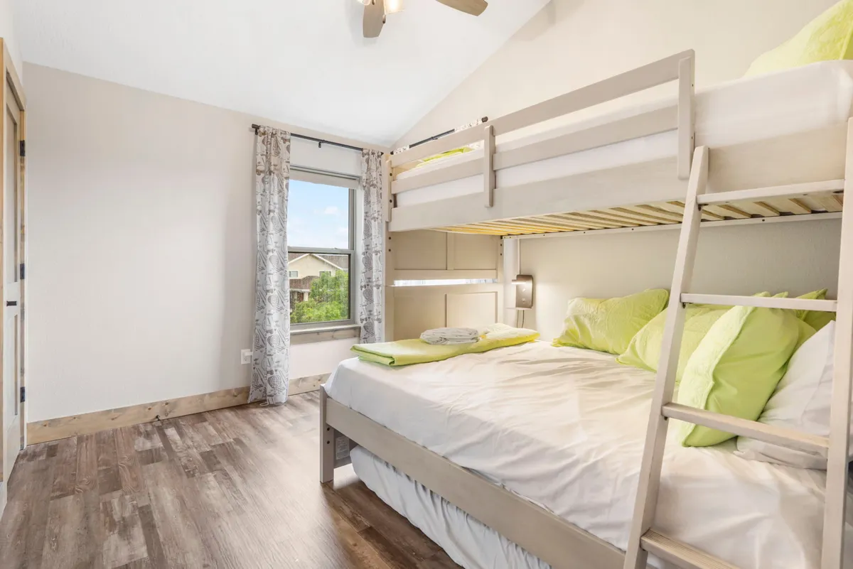 A bright and playful bunk room with a twin-over-full bunk bed, cheerful green pillows, a large window allowing in natural light, and decorative curtains. The space feels fun and inviting, perfect for kids or teens to relax and play.