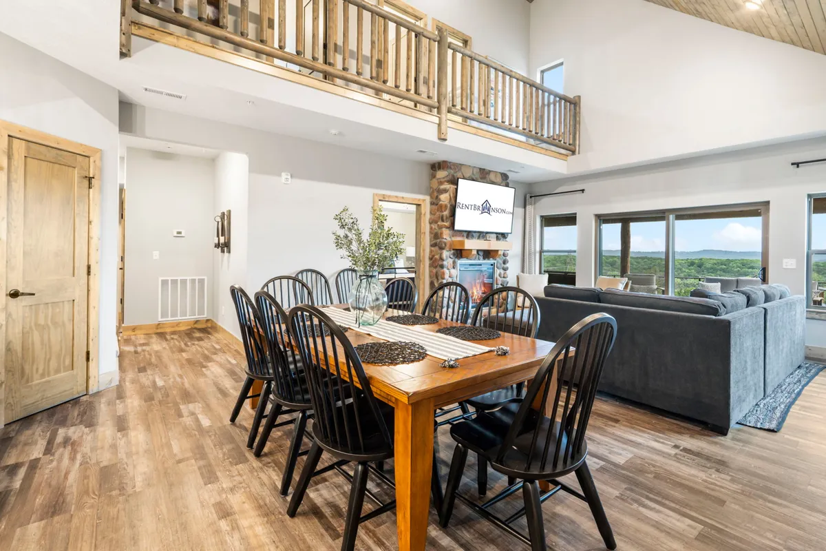 A cozy dining area anchored by a stone fireplace, with a large table set for gatherings. Expansive windows frame scenic views, and the atmosphere is warm and inviting, suggesting leisurely family meals and relaxed evenings.