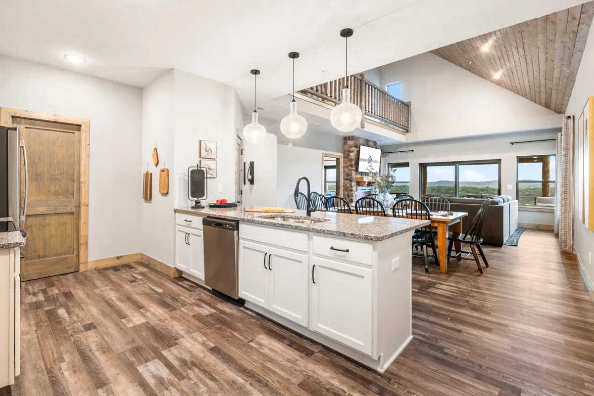 An expansive open-concept living space that seamlessly integrates the kitchen, dining area, and living room. The kitchen island serves as a hub for culinary activity, with large windows bringing in abundant natural light and offering views from both the sofa and dining table.