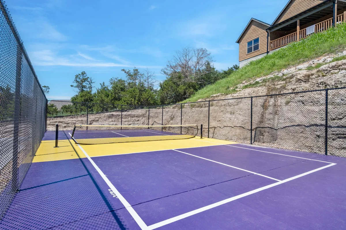 An active tennis court surrounded by lush greenery and a house in the background. Players are engaged in a match while others gather casually on the sidelines, creating an inviting atmosphere for recreation and socializing.