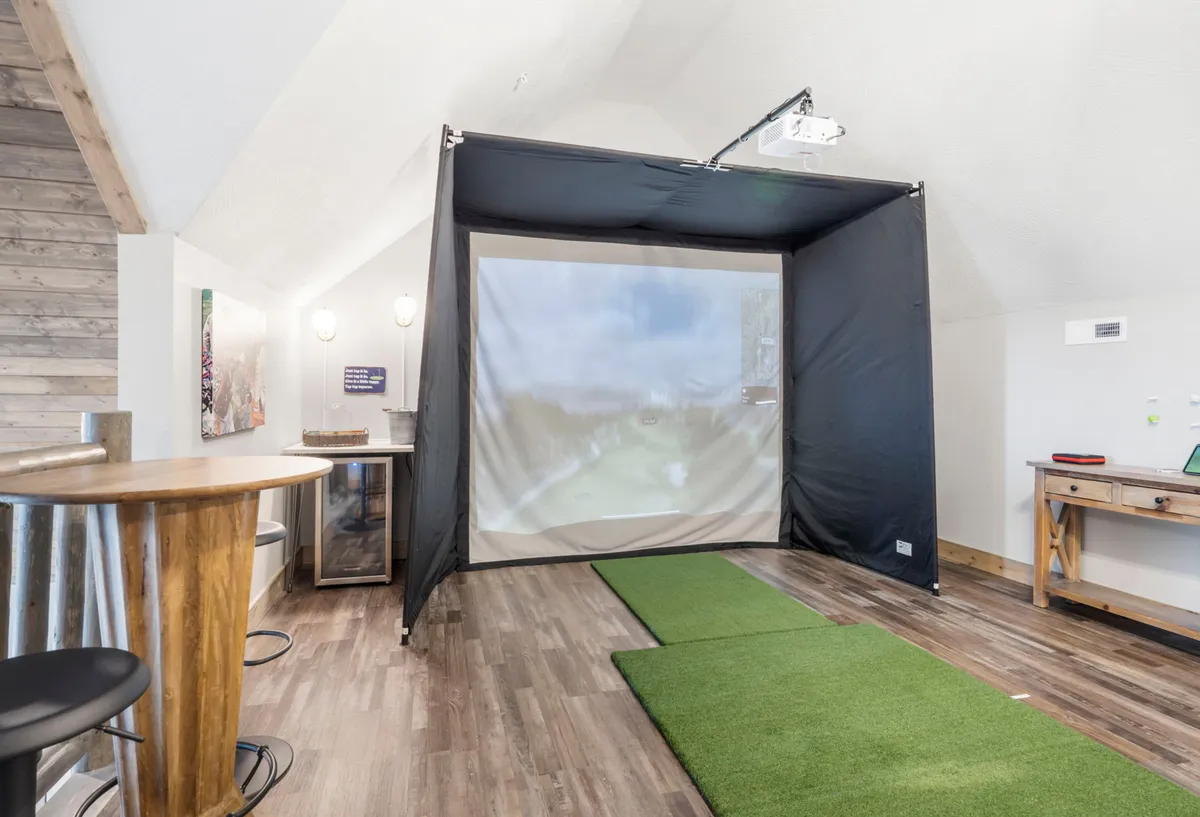 This image features a sleek indoor golf simulator setup with a large simulation screen, a realistic putting green, and an adjacent bar table, all within a comfortable upstairs recreation area. The scene is set for both serious golf practice and fun social gatherings, with the space designed for year-round enjoyment regardless of weather.