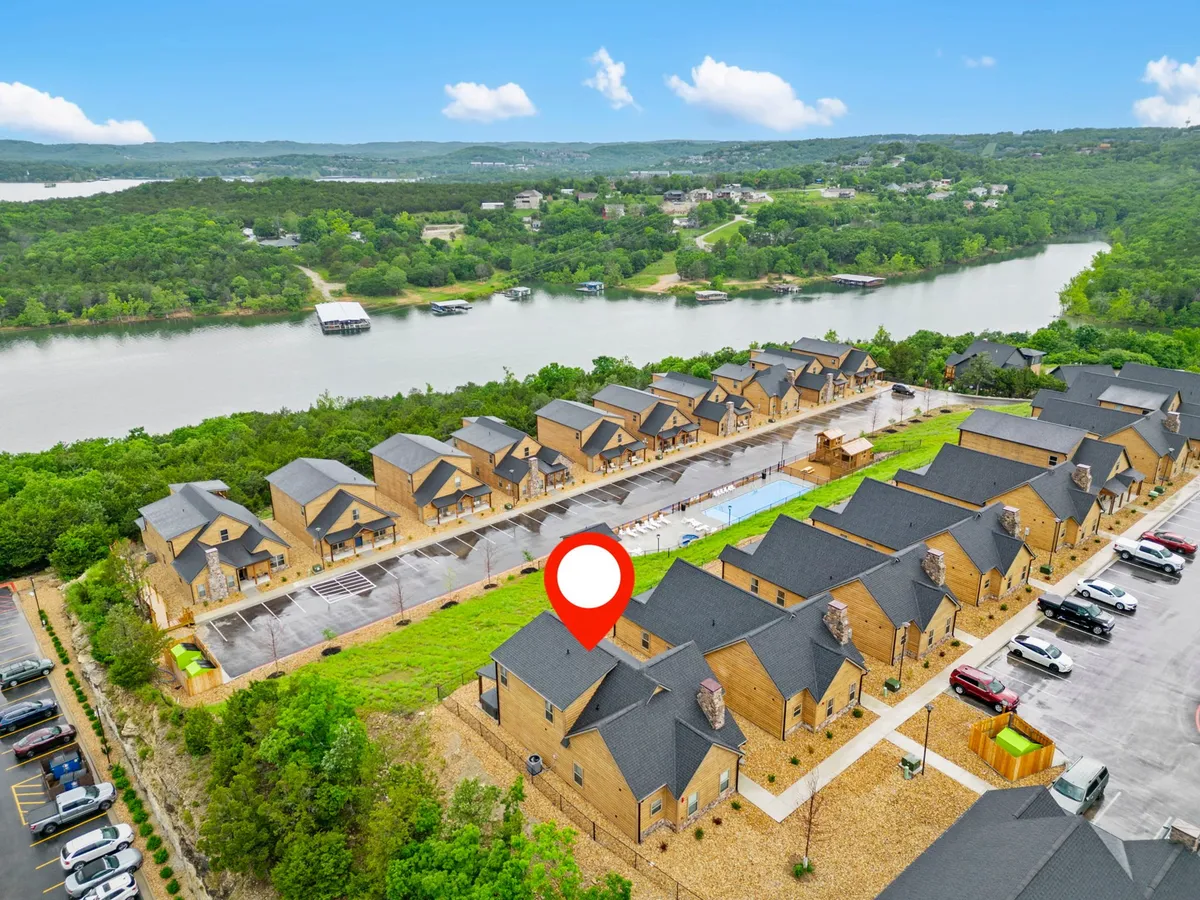 This image features a row of houses with a swimming pool in the foreground and a scenic river view beyond. The setting is outdoors, with visible community amenities like the pool, lounge chairs, and the natural beauty of the river and trees, evoking a sense of relaxed community living close to nature and recreation.