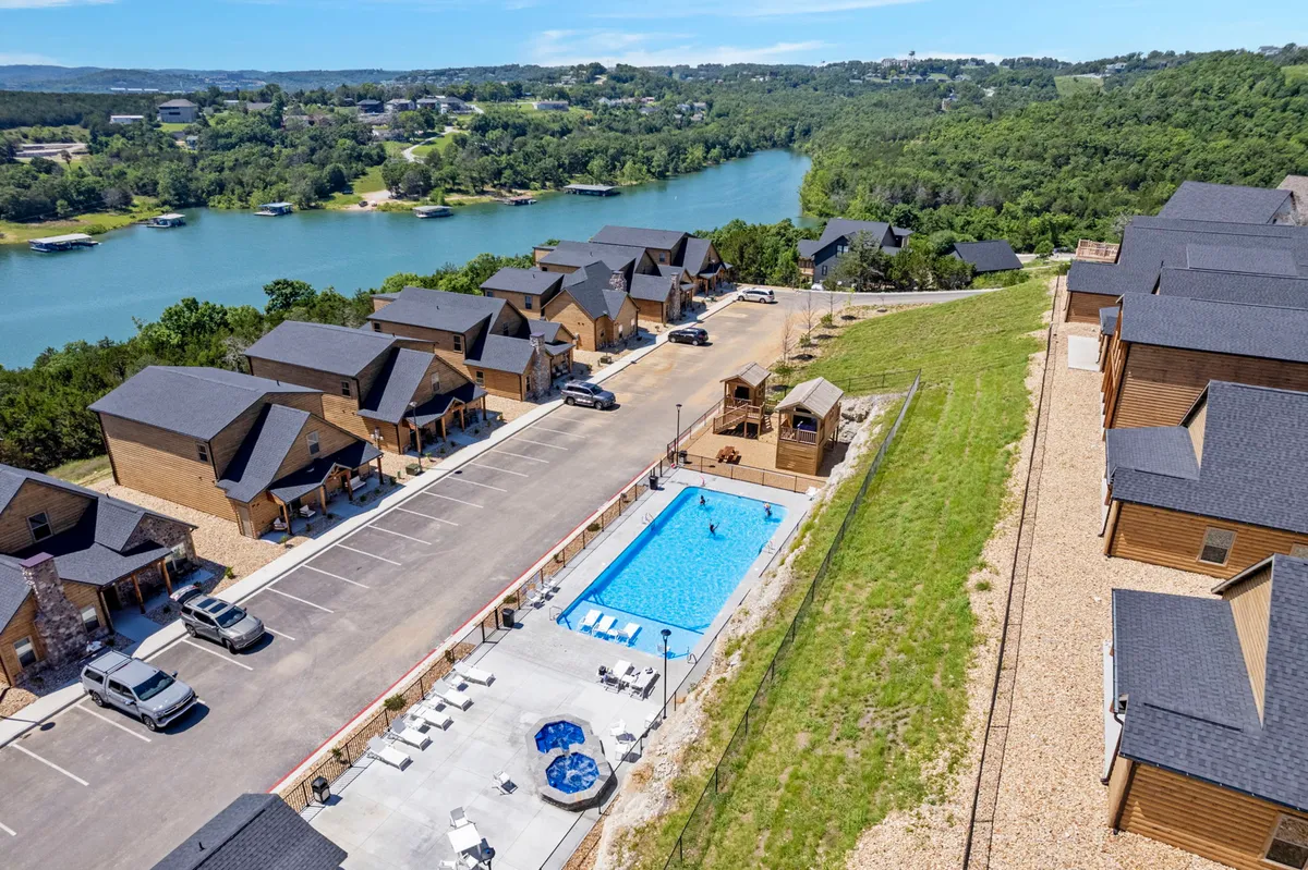 A spacious outdoor pool area featuring a sparkling swimming pool, comfortable lounge chairs, and a view of the lake framed by inviting wooden cabins. The setting suggests both active fun and peaceful relaxation, with the water reflecting the sky and the surrounding greenery enhancing the sense of escape.